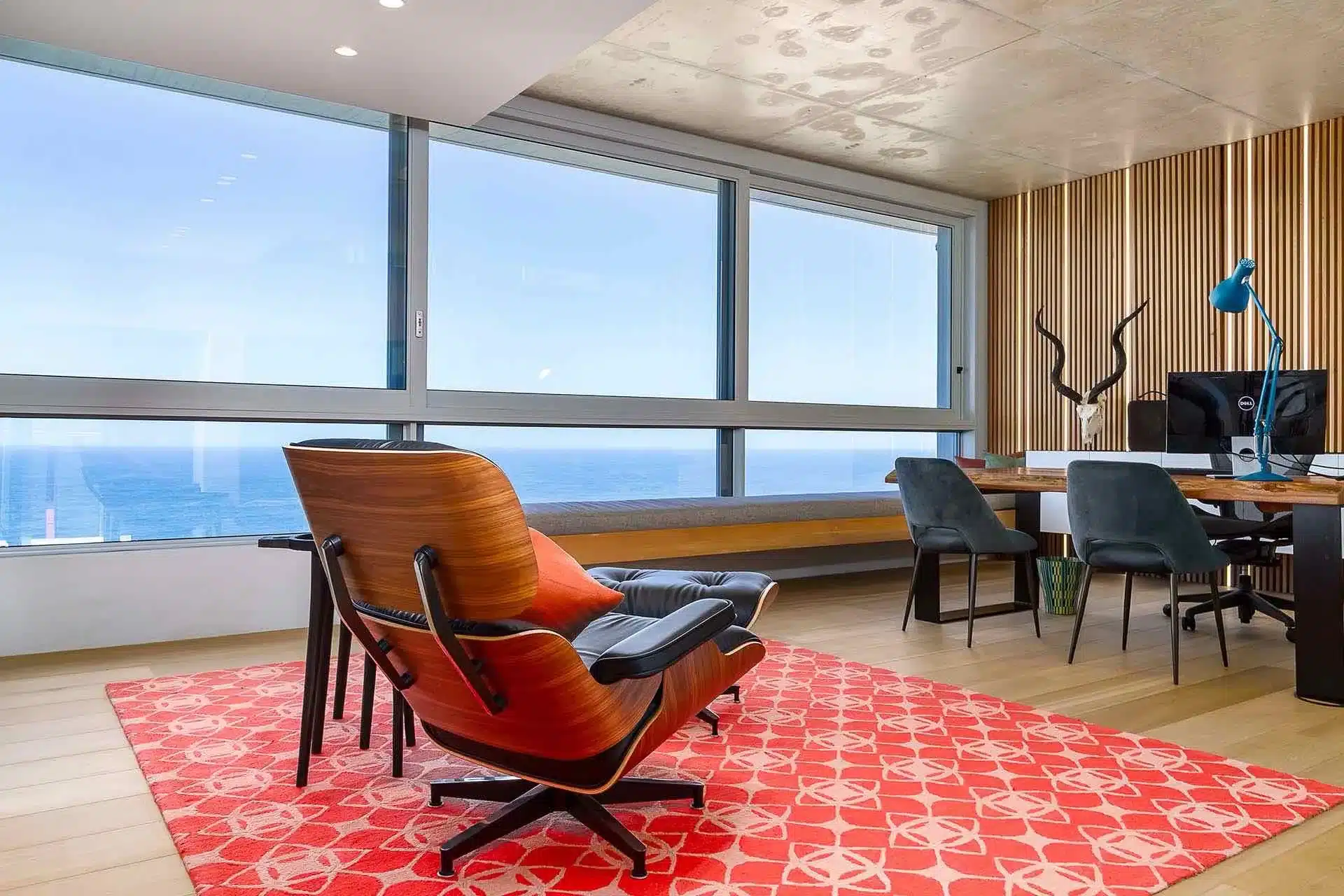 Modern office with large windows, wooden lounge chair, red patterned rug, desk with computer, blue chairs, and coastal view—perfect for managing concierge travel services or luxury villa rental bookings.