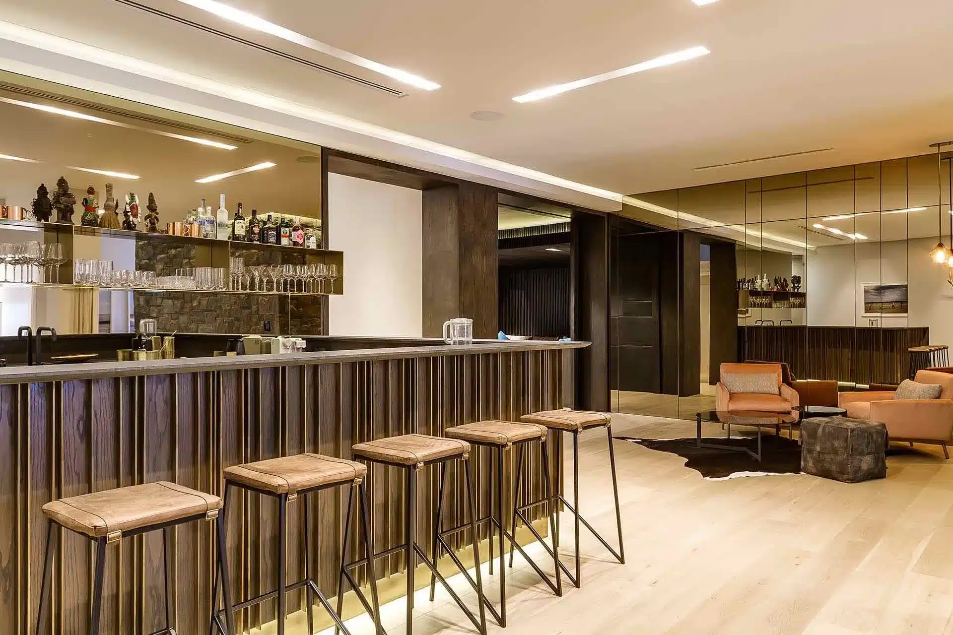 Modern bar area with wooden stools, mirrored walls, and bottle-lined shelves—perfect for relaxing in a luxury vacation rental after a day of bespoke concierge travel services.