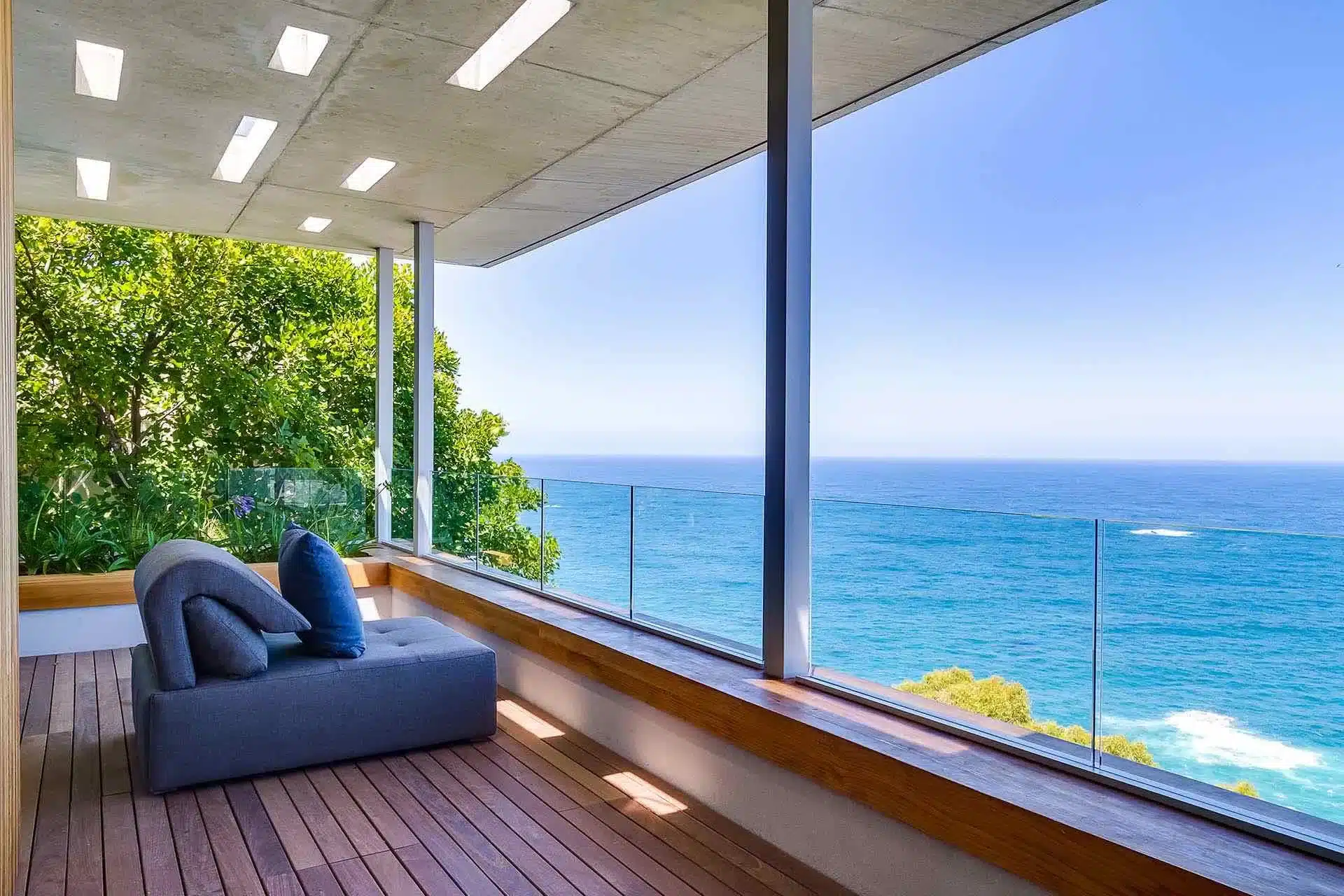 A modern balcony with a gray sofa and blue cushions overlooks the ocean—perfect for a luxury villa rental—with glass railings, wooden flooring, and lush greenery on the left.