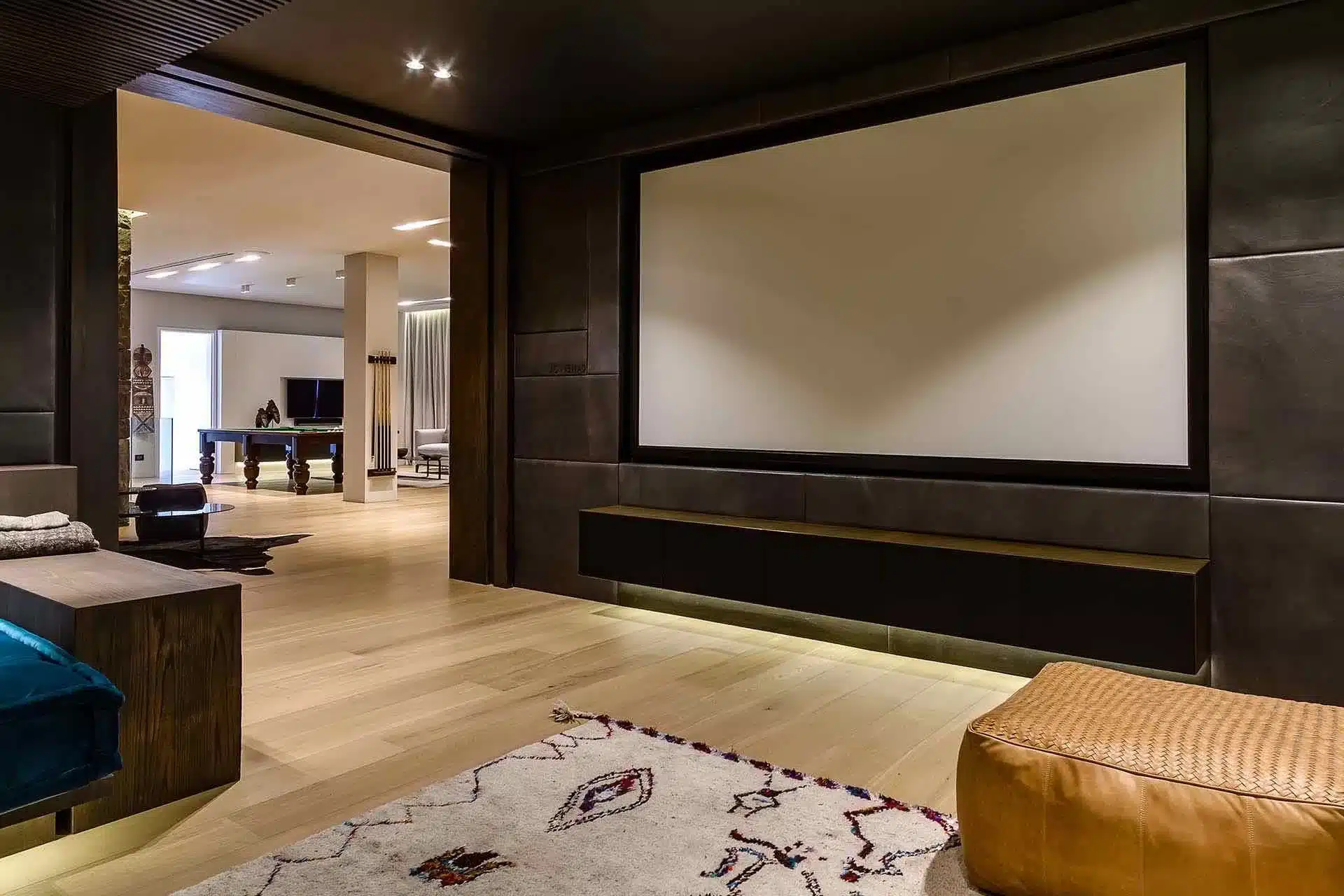 Modern home theater in a luxury villa rental features cozy seating, wood flooring, and a blank screen, leading to a bright room with pool table and TV for an elevated stay.