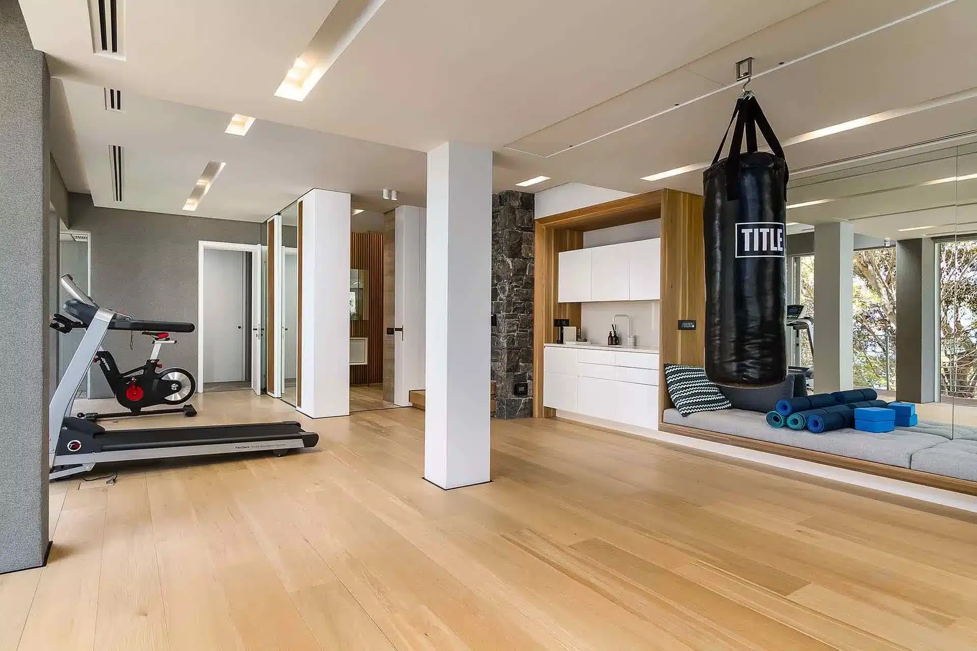 Modern home gym with a treadmill, punching bag, yoga mats, and cushioned lounge, perfect for a luxury vacation rental with wooden floors and large windows.