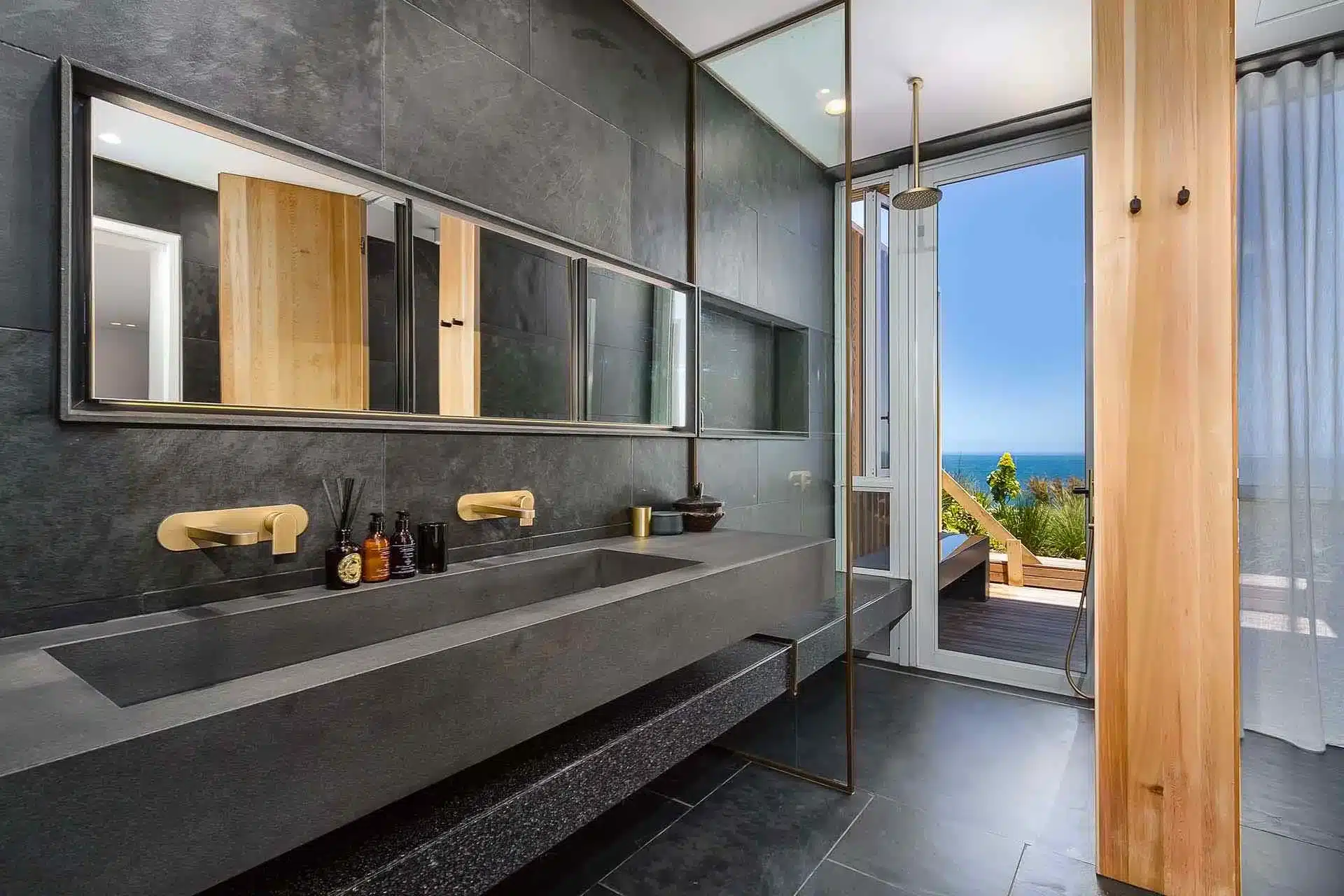 Modern bathroom with dark tiles, double sink, gold fixtures, and open shower area leading to a balcony—perfect for a luxury villa rental with stunning ocean views.