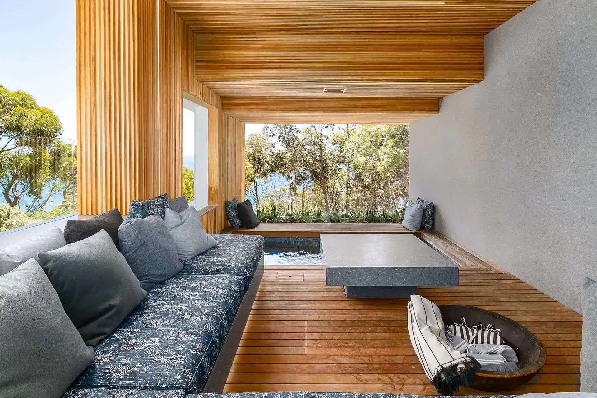 Modern outdoor lounge with wood paneling, gray sectional, low table, dog bed, and small pool—perfect for a luxury villa rental overlooking trees and water through open windows.