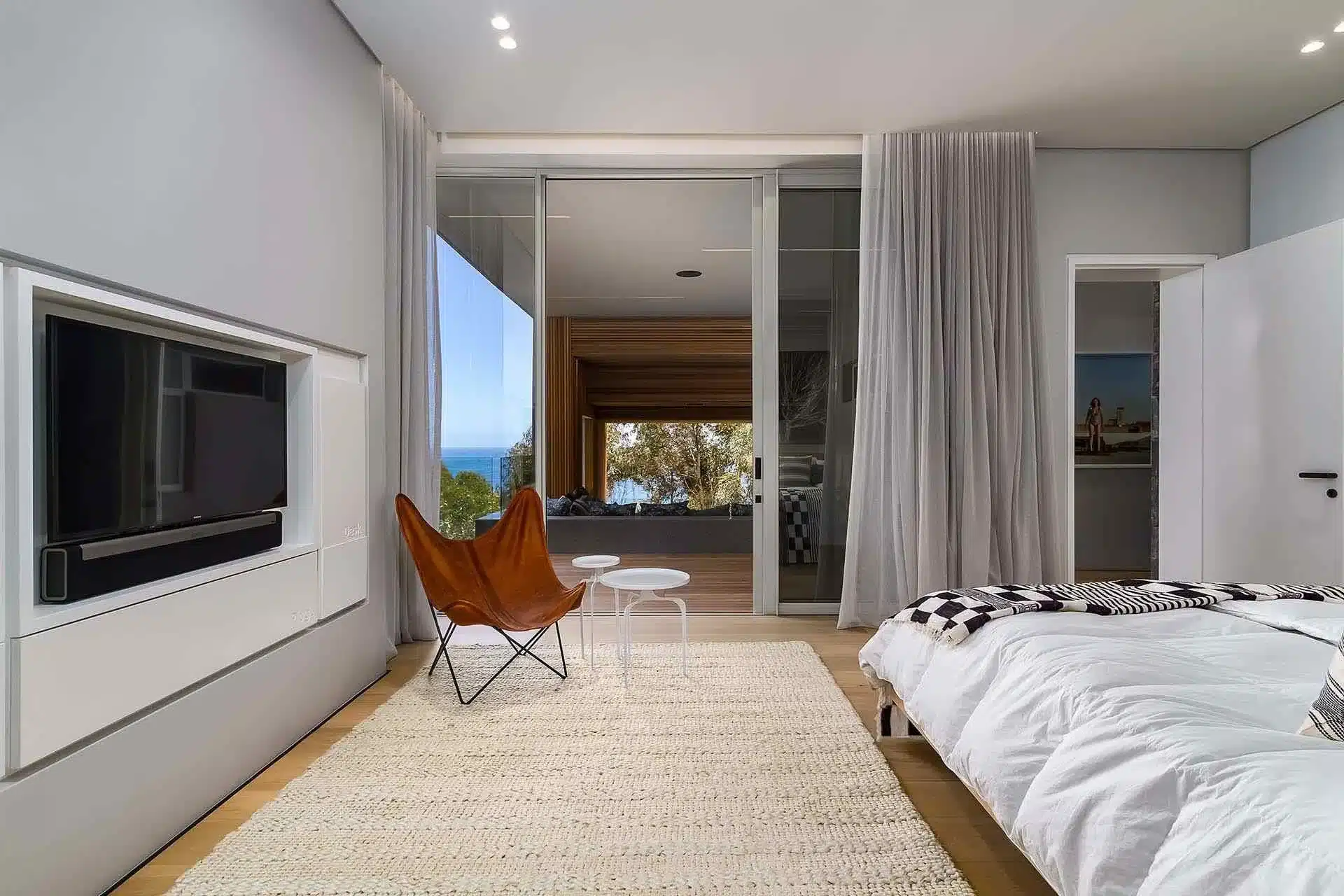Minimalist bedroom with a white bed, butterfly chair, and large glass doors opening to a patio—perfect for your next luxury villa rental or luxury vacation rental escape.