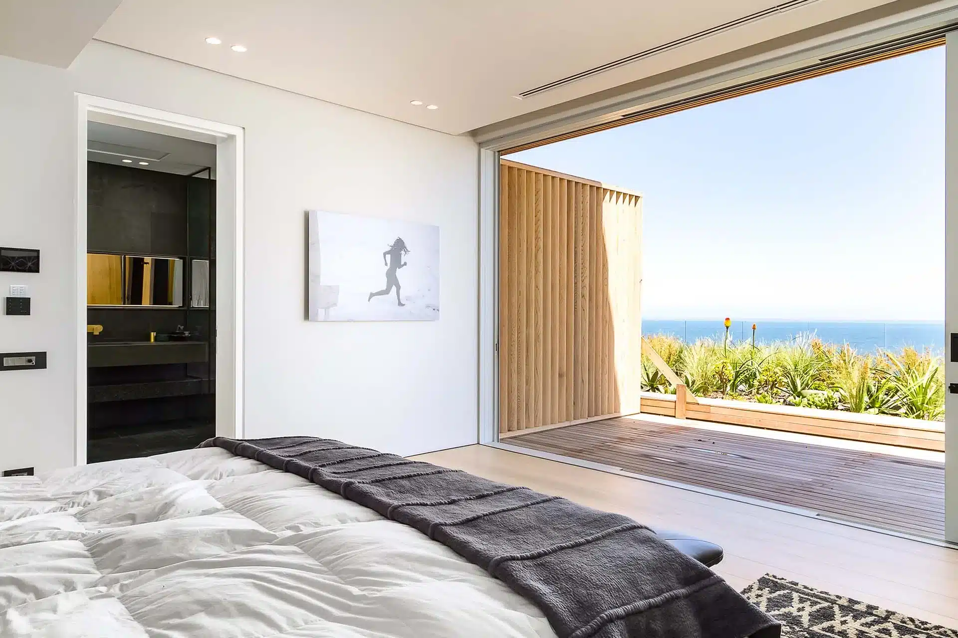 Modern bedroom with white walls and bedding, a dark gray throw, ocean-view deck, and bathroom entrance—ideal for a luxury vacation rental with available concierge travel services.