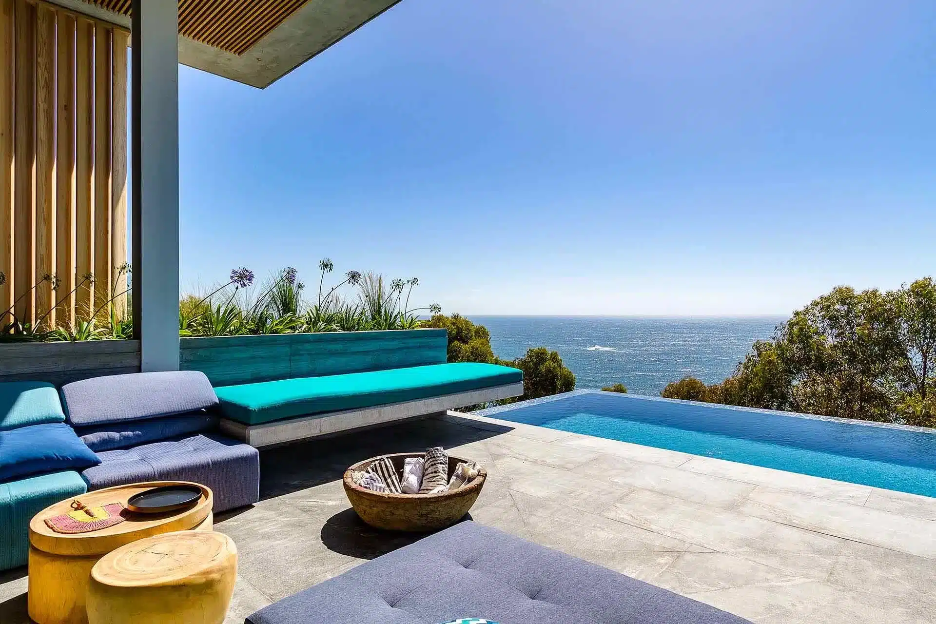 Modern patio at a luxury villa rental with blue lounge chairs, an infinity pool, wooden tables, and scenic ocean views. Trees surround this tranquil setting.