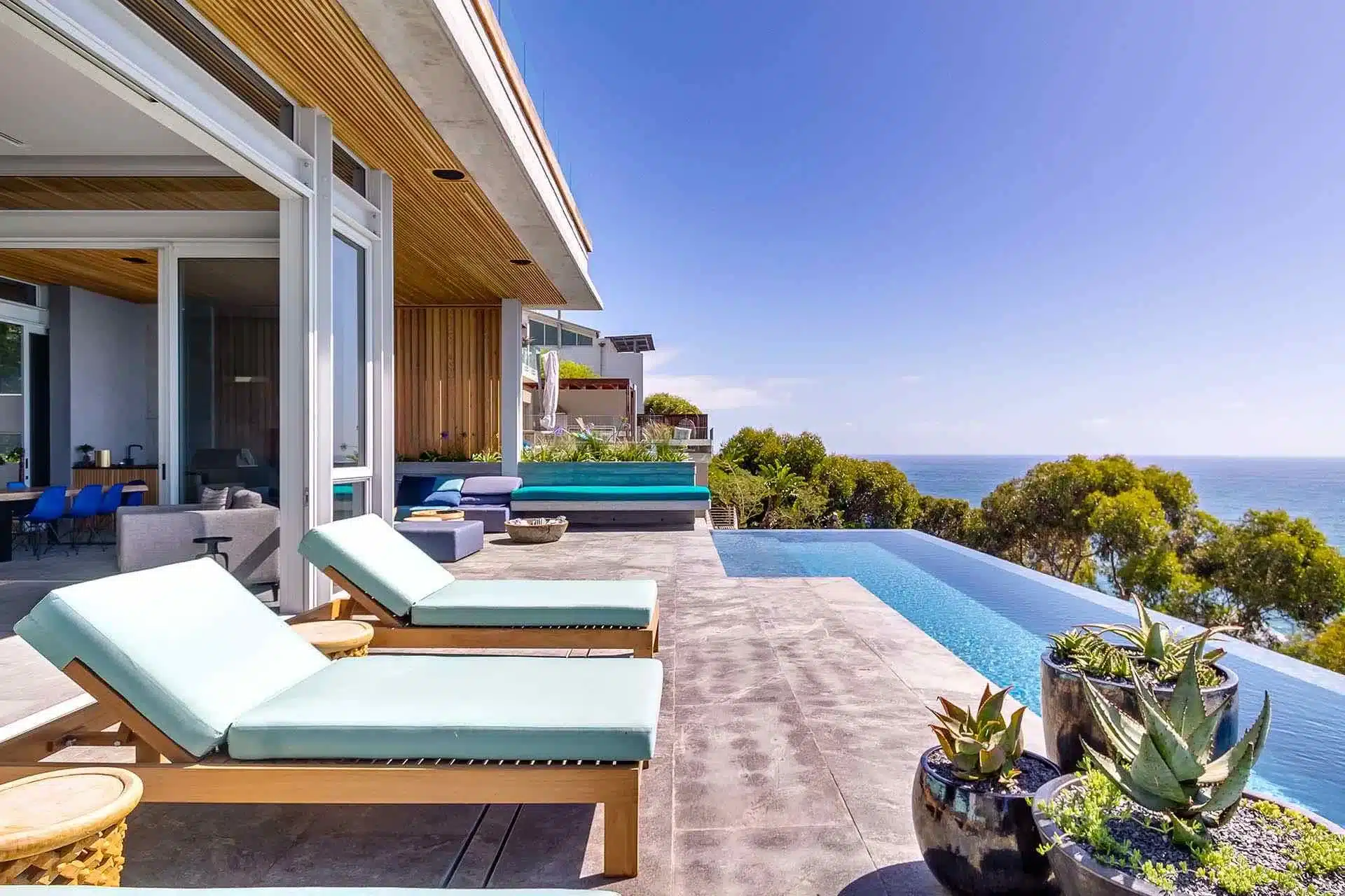 Modern house with open patio, two lounge chairs by an infinity pool, potted plants, ocean view, and a clear blue sky—perfect for a luxury villa rental.