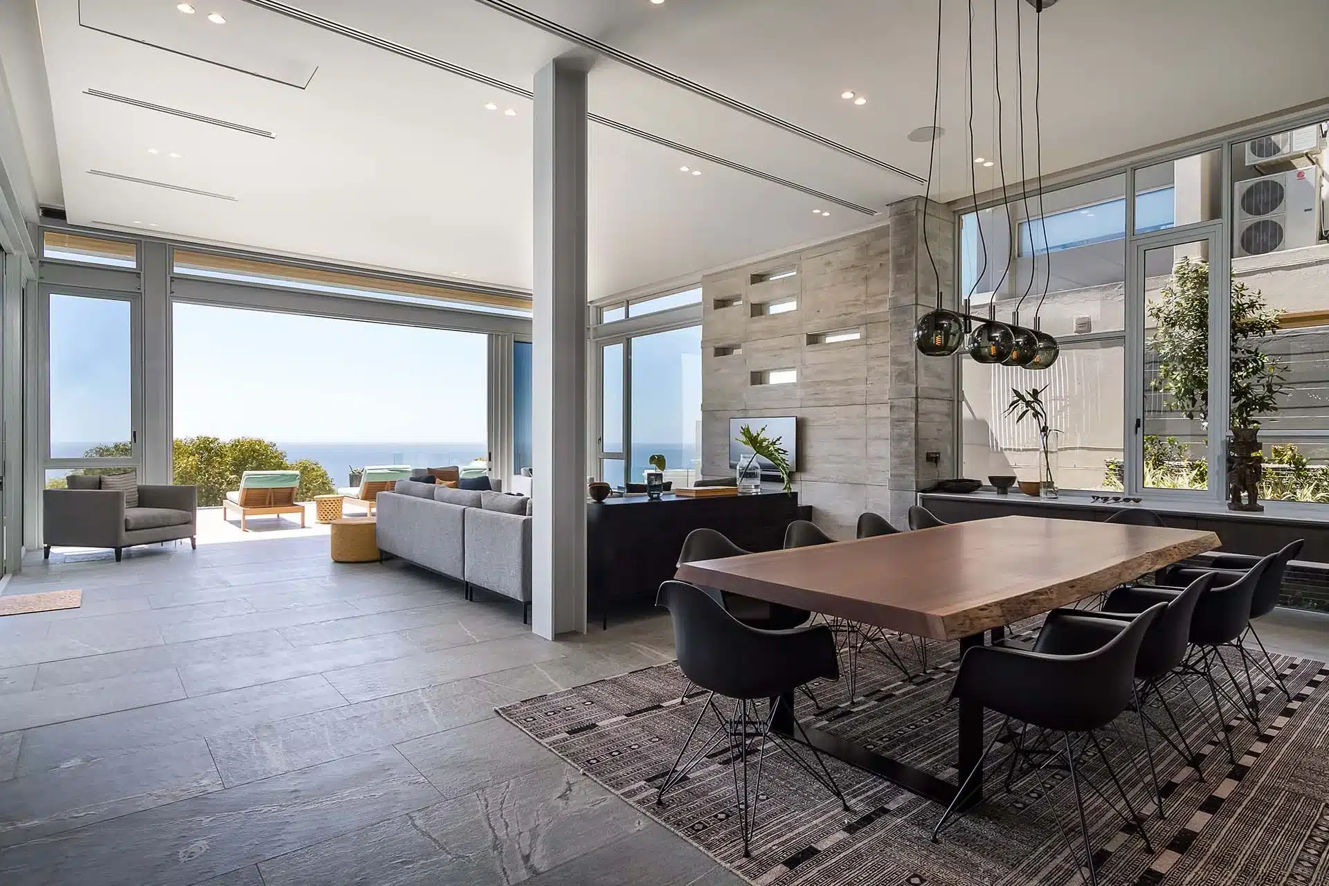 Modern open-plan living and dining area with large windows, ocean view, wooden table, black chairs, contemporary decor—perfect for a luxury villa rental experience.