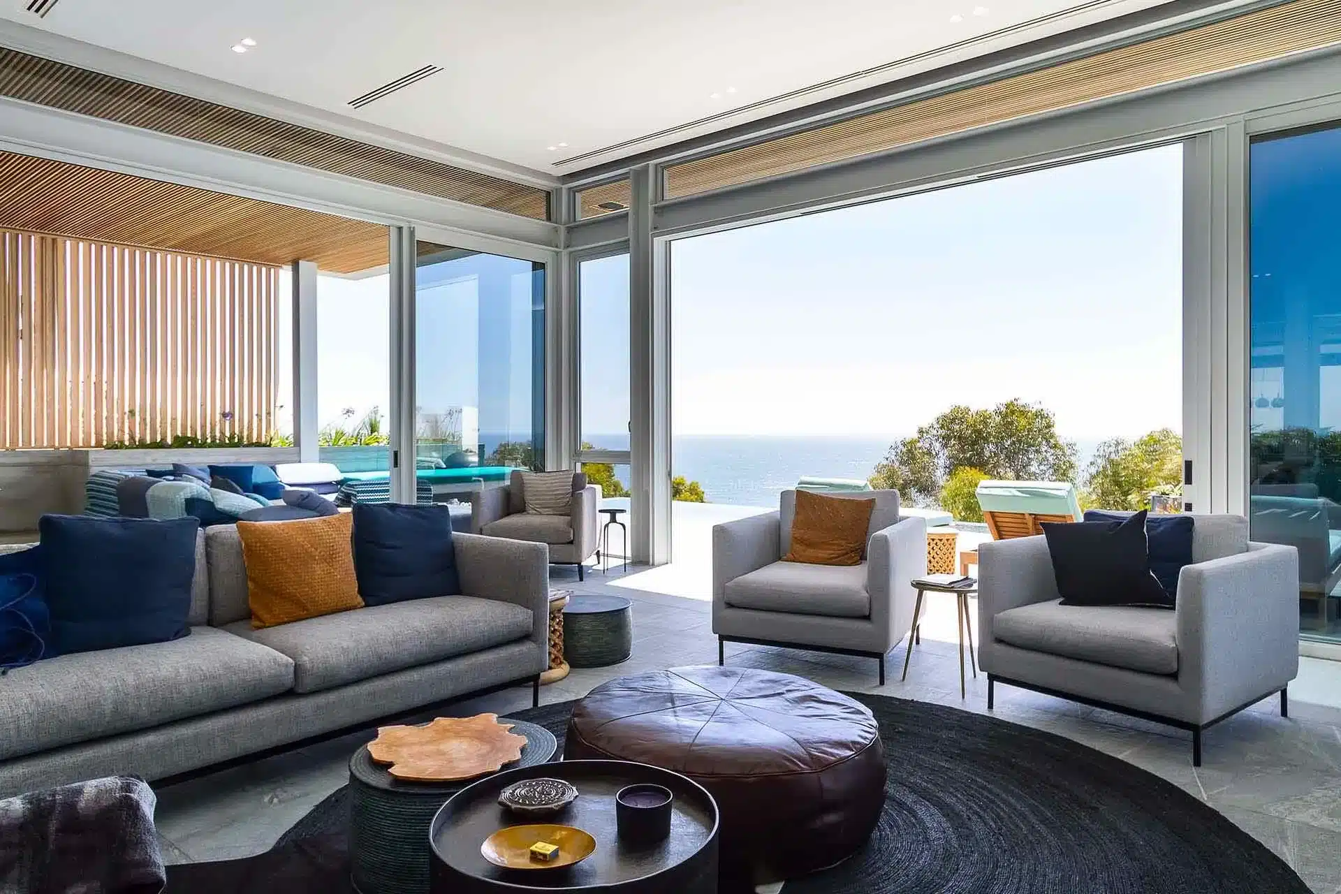 Modern living room with gray sofas, armchairs, and a round ottoman, perfect for a luxury villa rental, featuring large glass doors opening to a patio and ocean view.