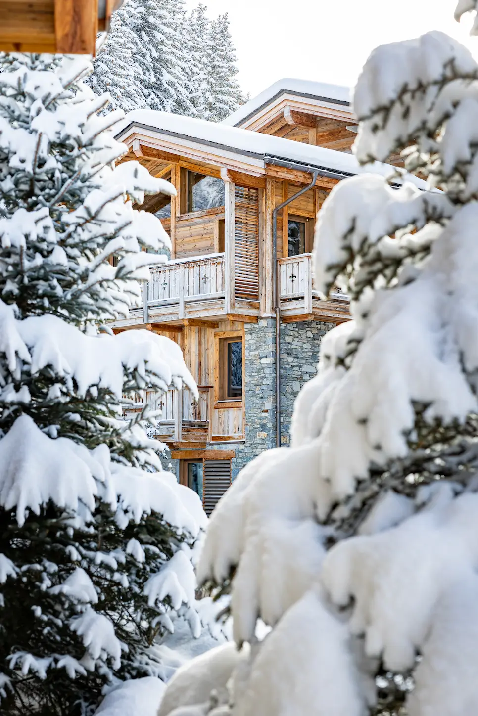 A wooden chalet with stone walls is nestled among snow-covered pine trees in a winter landscape.