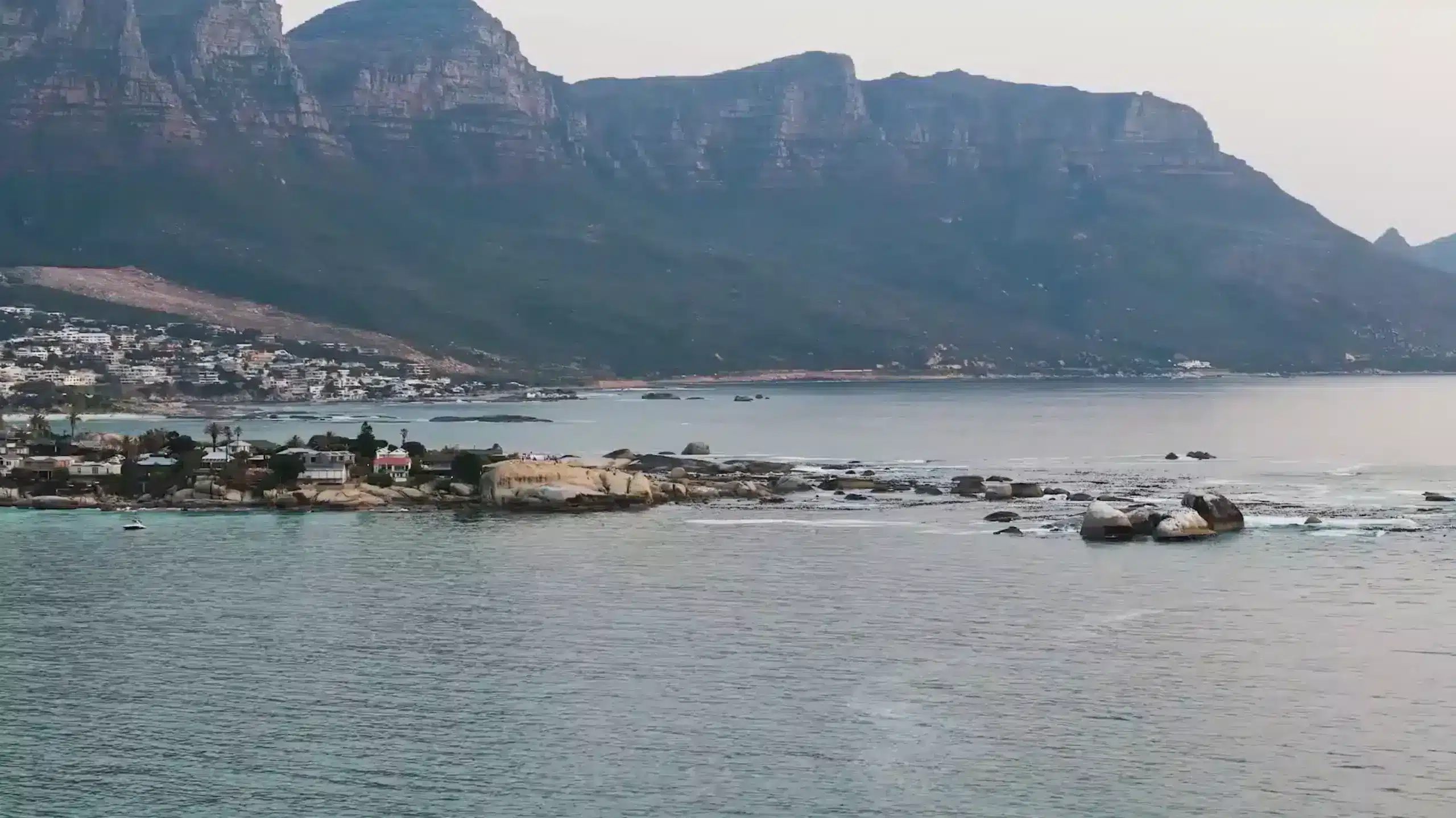Coastal scene with calm sea, large rocks, and distant mountains. Luxury villa rentals line the shoreline beneath a cloudy sky, offering serene views and refined comfort.