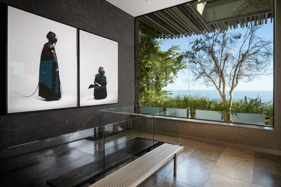A modern interior in Cape Town with two abstract portraits, a glass display case, a bench, and a large window overlooking trees and the ocean—perfect for private art tours.