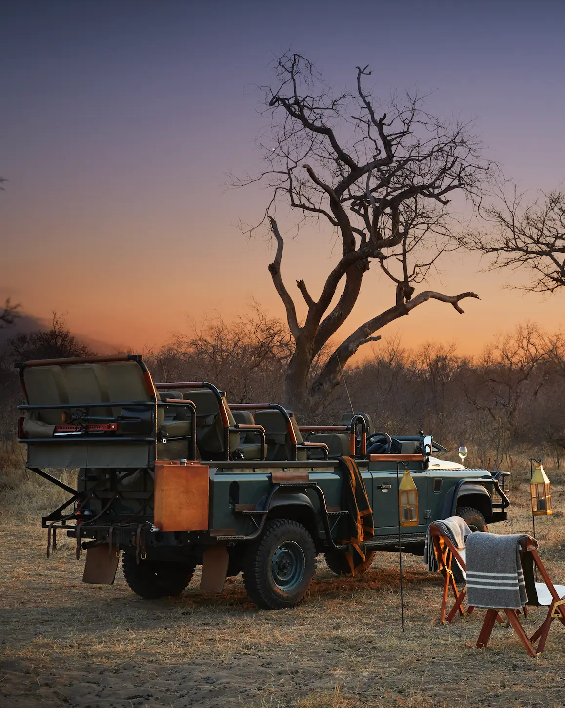 An open safari vehicle parked near a large leafless tree at sunset, with lanterns, a table, and chairs set up beside it in the wilderness.