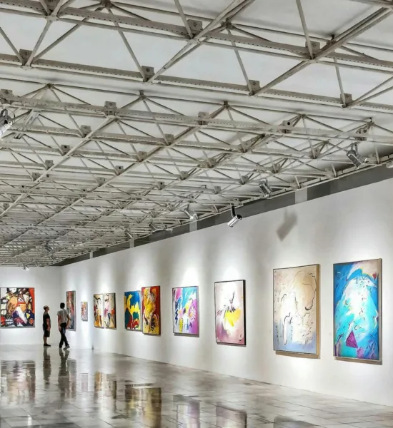 A spacious art gallery in Cape Town with white walls displays colorful paintings. Three people stroll the polished floor, enjoying the feel of a luxury event.